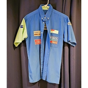 May include: Blue short-sleeve button-down shirt with lime green sleeves. The shirt features patches with the words "BARCIA", "Philips Car Stereo", "SASOL JORDAN", and "Yamaha". The shirt is hanging on a white hanger.