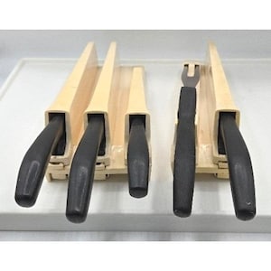 Può includere: Un set di cinque utensili da cucina con manici neri, composto da quattro coltelli e una forchetta, ciascuno inserito in fessure individuali all'interno di un blocco portaoggetti in plastica beige chiaro, disposti in due sezioni separate.