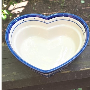 Puede incluir: Un cuenco de cerámica en forma de corazón con un interior de color crema y un borde azul intenso. El borde presenta un patrón decorativo de pequeños puntos azules espaciados uniformemente a lo largo del borde interior de este plato.