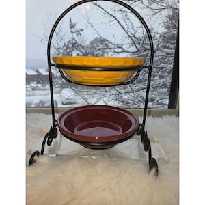May include: A two-tiered serving stand with a black wrought iron frame. The top bowl is yellow, and the bottom bowl is burgundy. The stand is on a clear acrylic base, with a snowy winter scene visible through the window in the background.