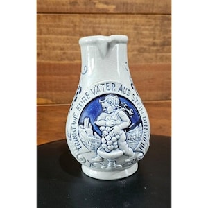 May include: A white and blue ceramic pitcher with a spout and a decorative relief. The relief features a cherub holding grapes, surrounded by text. The pitcher is likely for serving beverages.