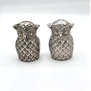 May include: A pair of silver-colored owl-shaped salt and pepper shakers. The shakers have detailed feather and eye designs. Each shaker has a small hole on top for dispensing salt or pepper. The owls are standing upright.