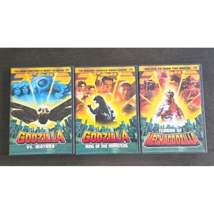 Puede incluir: Tres DVD de películas de Godzilla. Las portadas presentan imágenes de Godzilla, Mothra y Mechagodzilla, con texto que incluye "Godzilla vs. Mothra", "King of the Monsters" y "Terror of Mechagodzilla".