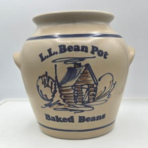 May include: Tan ceramic L.L. Bean Pot with blue accents. The pot features a cabin illustration and the words "L.L. Bean Pot" and "Baked Beans" in blue. It has two small handles and a wide opening.