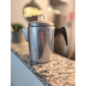 May include: A vintage aluminium coffee percolator with a black handle and a clear glass knob on the lid. The percolator is on a speckled countertop. The overall theme is kitchenware.