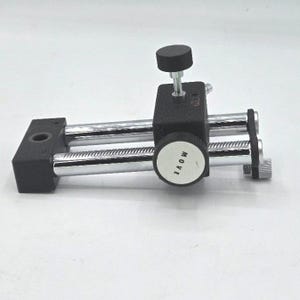 May include: A black and silver mechanical device with a knob labeled "MOVE". The device has two parallel silver rods and a black rectangular base. A small black knob sits atop a black rectangular block.