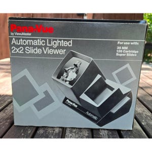 May include: A black and gray product box for a Pana-Vue Automatic Lighted 2x2 Slide Viewer. The box features the product name, text indicating compatibility with 35mm and 126 cartridge super slides, and a graphic illustration of the slide viewer.