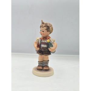 May include: A ceramic figurine of a young boy in a traditional outfit. The figurine features a brown hairstyle, a red bow tie, and a detailed outfit with brown shorts and a vest. The boy is holding items in his hands and stands on a round base.