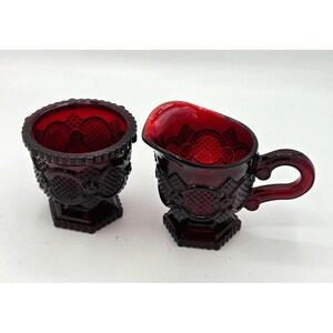 May include: A ruby red glass creamer and sugar bowl set. The creamer has a handle and a spout. Both pieces have a detailed pattern of circles and diamond shapes. The sugar bowl is round with a scalloped rim. The set sits on a white surface.