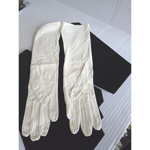 May include: A pair of off-white, long leather gloves. The gloves are laid out on a black surface, with the fingers pointing down. The gloves appear to be made of a soft, supple leather. The gloves are likely for formal wear.