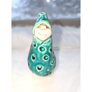 May include: A ceramic gnome figurine with a teal green body and a white face. The gnome's body has a series of circular indentations. The gnome wears a pointed hat and has a smiling expression.