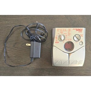 Puede incluir: Un pedal de efectos Zoom 506II Bass plateado con un adaptador de corriente y cable negros. El pedal tiene varias perillas y botones, y una pequeña pantalla roja. El logotipo de Zoom es visible.