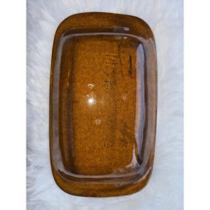 May include: A rectangular, brown ceramic serving dish with a glossy finish. The dish has rounded corners and a slightly raised edge. The bottom of the dish has embossed text and markings. The dish is set against a white, fluffy background.