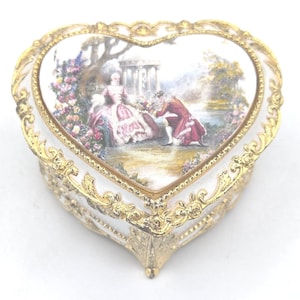 May include: A heart-shaped trinket box with a white base and gold filigree trim. The lid depicts a painted scene of a couple in period dress in a garden. The box has an ornate, decorative design.