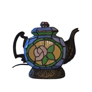 Könnte beinhalten: Eine Tiffany-Teekannenlampe mit floralem Design. Die Lampe hat eine rosa Rose in der Mitte, umgeben von grünen Blättern und gelben und blauen Glaspaneelen. Der Sockel und der Griff sind dunkelbraun.