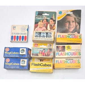 May include: A collection of vintage flash cubes and flash bulbs in their original packaging. Brands include Westinghouse, GE, and Sylvania. The boxes feature colorful graphics and text, including "Flash Cubes" and "MagiCubes".