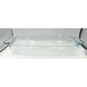 Puede incluir: Una fuente de horno rectangular de vidrio transparente con esquinas redondeadas y asas. La fuente está vacía y se encuentra sobre una superficie blanca. El vidrio es transparente.
