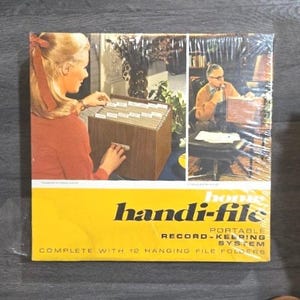 May include: A vintage "homes handi-file" record-keeping system, complete with 12 hanging file folders. The box is yellow with black text and an image of a woman and a man. The file box is brown.