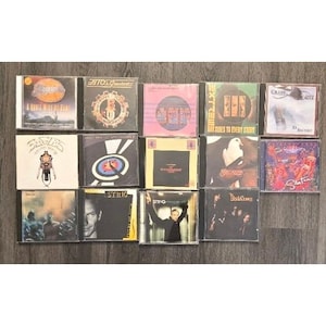 Op de afbeelding: Een verzameling van veertien muziek-cd's in plastic doosjes, gerangschikt in drie rijen. De albums bevatten diverse hoesontwerpen, waaronder titels als America, BTO's Greatest, Extreme, Eagles, Santana en Sting, met uiteenlopende grafische stijlen.