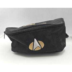 May include: A black waist bag featuring a zippered top closure and an embroidered Star Trek Starfleet delta insignia on the front. The emblem consists of a gold oval background with a silver triangular shape centered within the design.