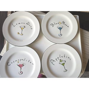 Puede incluir: Cuatro platos de cerámica blanca con bordes negros, cada uno con un diseño de cóctel diferente. Los platos están etiquetados como "Lemon Drop", "Blue Moon", "Cosmopolitan" y "Appletini", con ilustraciones.