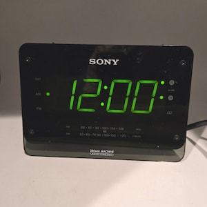Puede incluir: Reloj despertador digital Sony Dream Machine negro que muestra las 12:00 en dígitos verdes. Cuenta con indicadores de frecuencia de radio FM y AM y ajustes de alarma. El reloj es rectangular con esquinas redondeadas.