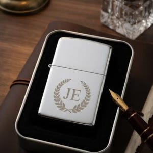 May include: A rectangular silver-toned metal lighter with a polished finish, featuring an engraved design of the letters JE centered within a circular laurel wreath. The lighter is presented inside a black-lined metal storage case.