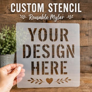 May include: A square, semi-transparent Mylar stencil featuring the cut-out text "YOUR DESIGN HERE" in a bold, sans-serif font. Below the text is a small heart icon flanked by two decorative leaf sprigs. The stencil is held against a wooden background.
