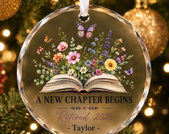 May include: A round, clear glass ornament featuring an open book with a colorful floral arrangement emerging from the pages. Butterflies are depicted above the flowers. The text reads A NEW CHAPTER BEGINS, Retired 2026, and - Taylor -.