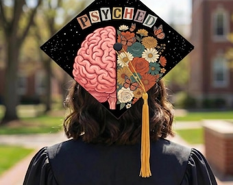 May include: A black graduation cap with the word "PSYCHED" in colorful letters. The cap features a brain illustration on the left and a floral design with butterflies on the right. A gold tassel hangs from the cap.