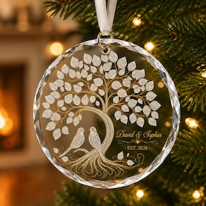 May include: A circular clear glass ornament with faceted edges, featuring a gold-toned etched design of a tree with two birds perched on its roots. The text "David & Sophia" and "EST. 2026" is engraved on the right side. A white ribbon is attached.