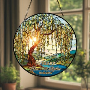 Op de afbeelding: Rond gebrandschilderd glazen raam met een treurwilg en een zonsondergang. Het kunstwerk bevat de tekst "10 JAAR", de namen "David & Sophia" en de datum "10.08.2015". Het raam is zwart omlijst en hangt aan een ketting.