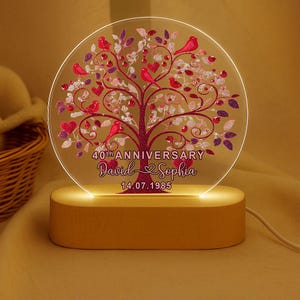 Peut inclure: Veilleuse LED personnalisée pour le 40e anniversaire. Le disque en acrylique transparent présente un motif d'arbre avec des oiseaux, des cœurs et des feuilles rouges. Les noms "David & Sophia" et la date "14.07.1985" sont affichés en dessous. La base est en bois.