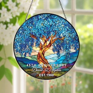 Op de afbeelding: Een rond gebrandschilderd glazen raam met een boom met blauwe bladeren en een bruine stam. De tekst "45th Anniversary David & Sophia EST.1980" staat onderaan. Het raam hangt aan een ketting.