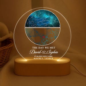 May include: A personalised night light with a clear acrylic disc showing a celestial map and a city map. The text "THE DAY WE MET" and names "David & Sophia" are displayed. The light sits on a wooden base.