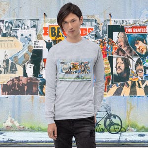 May include: A grey long-sleeved shirt featuring a colourful graphic print of a street scene with a bicycle. The graphic print is a collage of images of the Beatles.