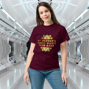 May include: A maroon t-shirt featuring a gold graphic of the text "Sgt. Pepper's Lonely Hearts Club Band" with a decorative design.