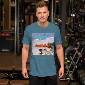 May include: A teal blue t-shirt with a graphic print of a mermaid on a beach. The graphic includes the word "Splash" in a blue font.