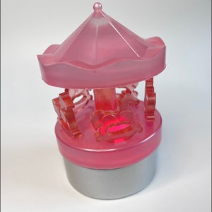 Puede incluir: Un carrusel de plástico rosa con una base iluminada y un fondo plateado. El carrusel tiene un techo rosa y cuatro caballos.