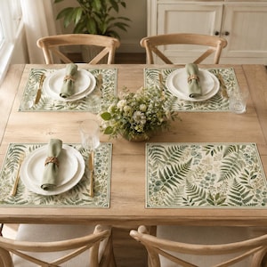 Può includere: Un tavolo da pranzo in legno apparecchiato per quattro persone con tovagliette rettangolari caratterizzate da un motivo a foglie di felce verdi e beige. Ogni posto tavola include un piatto in ceramica bianca, un tovagliolo di stoffa verde.
