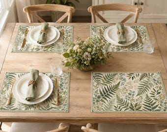 May include: A wooden dining table set for four with rectangular placemats featuring a green and beige fern leaf pattern. Each place setting includes a white ceramic plate, a light green cloth napkin with a woven ring, and gold cutlery.