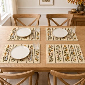 May include: Four rectangular placemats with a cream background and vertical stripes featuring autumn oak leaves, acorns, and berries. Each placemat holds a white round plate, a silver fork, a silver knife, and a clear glass on a wooden table.