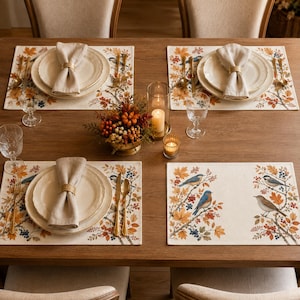 Puede incluir: Una mesa de comedor de madera preparada para cuatro personas con manteles individuales rectangulares que presentan ilustraciones de pájaros, hojas de otoño y bayas. Cada cubierto incluye un plato de cerámica blanca, una servilleta de lino beige con un anillo dorado y cubiertos dorados.
