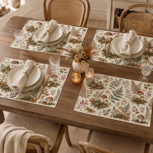 Pode incluir: Uma mesa de jantar posta para quatro pessoas com jogos americanos retangulares com uma estampa botânica de cogumelos, fetos e bagas. Cada lugar inclui um prato de cerâmica branca, um guardanapo de linho bege com um anel dourado, uma faca e um copo.