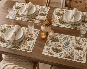 May include: A dining table set for four with rectangular placemats featuring a botanical print of mushrooms, ferns, and berries. Each setting includes a white ceramic plate, a beige linen napkin with a gold ring, a knife, and a glass.