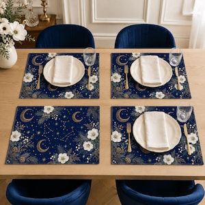 Peut inclure: Quatre sets de table rectangulaires sur fond bleu marine ornés de croissants de lune dorés, de fleurs blanches, de branches de pin et de constellations. Chaque couvert comprend une assiette blanche, une serviette pliée, une fourchette dorée et un verre.