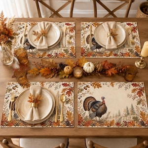 May include: A dining table setting featuring four rectangular placemats decorated with a turkey, autumn leaves, and acorns. Each place setting includes a cream-coloured plate, a linen napkin with a leaf napkin ring, and gold-toned cutlery.