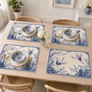 Pode incluir: Quatro jogos americanos retangulares com um padrão azul e branco de grous voadores e flores de cerejeira. Cada lugar inclui uma tigela de cerâmica branca, um prato branco, um copo transparente, uma colher e um garfo dourados.