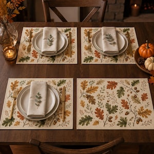 May include: Four rectangular placemats featuring a printed pattern of autumn oak leaves and acorns in shades of green, orange, and brown. Each setting includes a white ceramic plate, a beige linen napkin, a gold fork, and a gold knife.