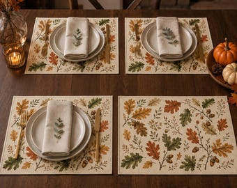 May include: Four rectangular placemats featuring a printed pattern of autumn oak leaves and acorns in shades of green, orange, and brown. Each setting includes a white ceramic plate, a beige linen napkin, a gold fork, and a gold knife.